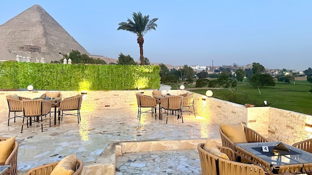 An outdoor seating area with wicker chairs and tables, a pyramid in the background, and a palm tree. - Golf Pyramids View (Giza)