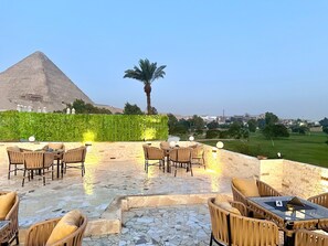 Un espace extérieur aménagé avec des chaises et des tables en osier, une pyramide en arrière-plan et un palmier.