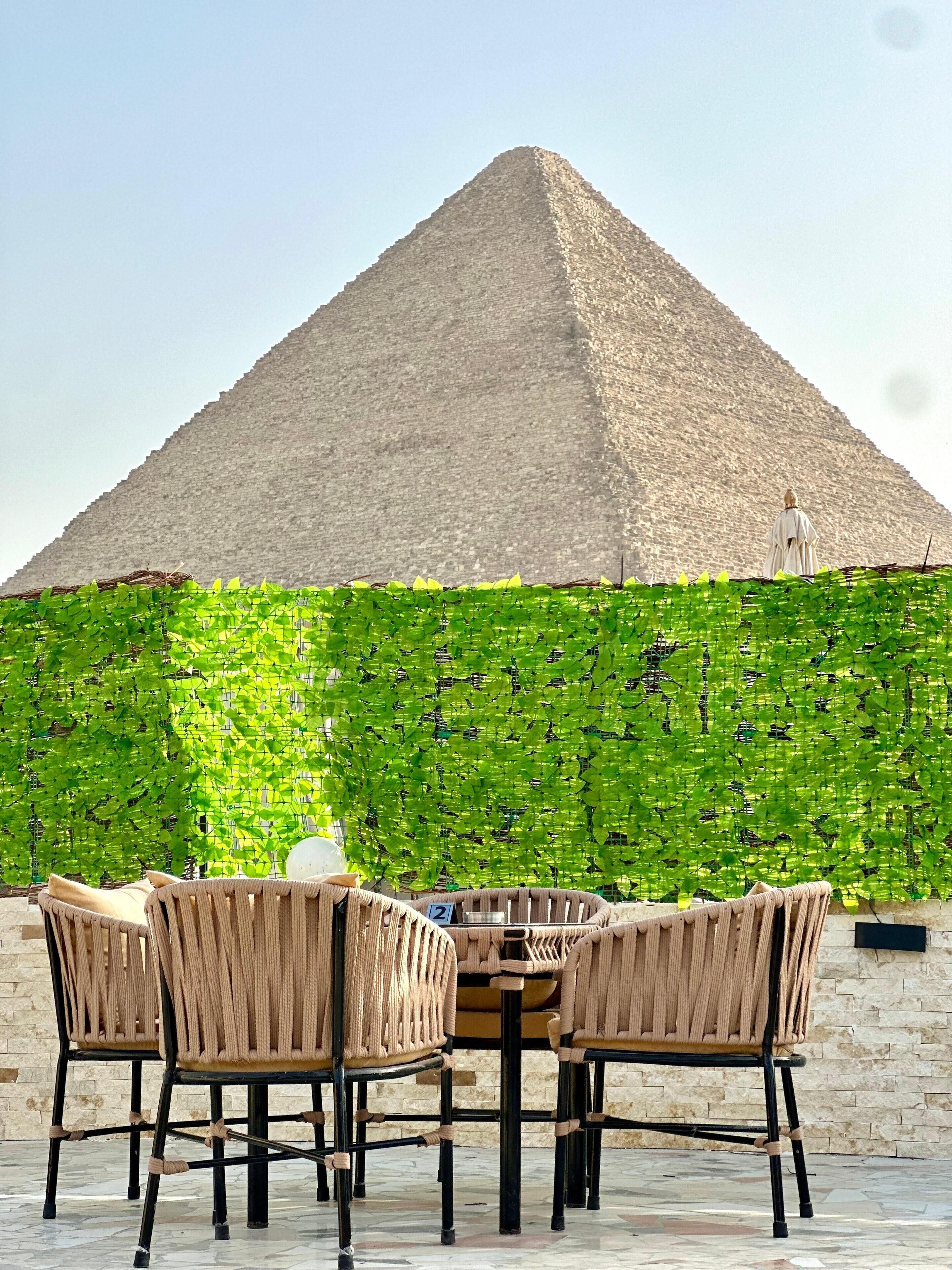 A dining area with wicker chairs and a table, a pyramid structure in the background, and a green leafy wall.