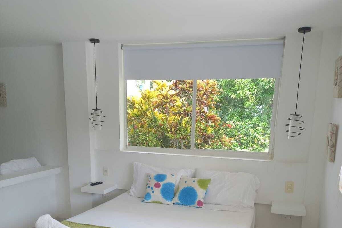 A bedroom with a bed, a window with a view of greenery, and a white color scheme.