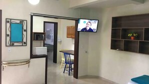 Un salon avec un téléviseur, un coin repas avec une table et des chaises, une cuisine à l’arrière-plan et un ventilateur de plafond.
