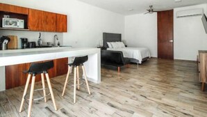 Une cuisine moderne avec des meubles en bois, un plan de travail blanc et un ventilateur de plafond. On aperçoit, au fond, un lit avec une tête de lit grise et une literie blanche.