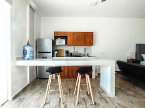 Una cocina moderna con encimera blanca, armarios de madera y refrigerador de acero inoxidable. Hay dos taburetes negros frente a la encimera.
