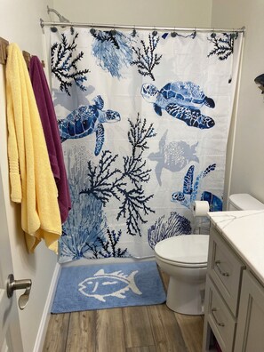 Ein Badezimmer mit Duschvorhang in Meereskugelfisch- und Korallenmuster, einem gelben Handtuch, einer blauen Fußmatte und einer weißen Toilette.