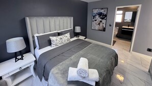A modern bedroom with a large bed, a bedside table with a lamp, a ceiling fan, and a bathroom visible through an open door. - Knightsbridge Luxury Apartments Century City by Vvip Bookings  (Cape Town)