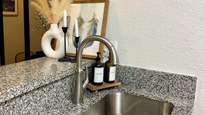 Un lavamanos de cocina con encimera de granito, un grifo y una pequeña bandeja de madera con dos botellas.