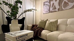 Un salón con una mesa de centro de madera oscura, un sofá blanco con cojines a rayas, una silla negra y una planta en un jarrón.