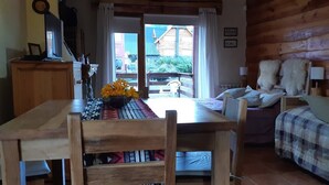 A cozy cabin interior with a wooden dining table, a bed with plaid bedding, and a view of a wooden house through a glass door. - Cerro Catedral excellent studio apartment (Cerro catedral,Río Negro Province)