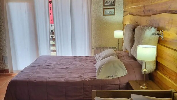 A cozy bedroom with a wooden headboard, a large bed with a pink bedspread, two bedside tables with lamps, and a view of the outside through curtains. - Cerro Catedral excellent studio apartment (Cerro catedral,Río Negro Province)