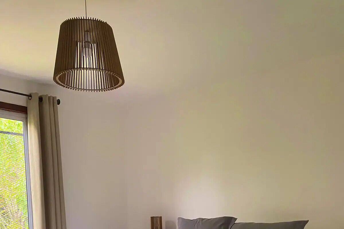 Ein ordentlich bezogenes Bett mit weißer Daunendecke und grauen Kissen, ein schwarzer Nachttisch und eine gewebte Pendelleuchte.