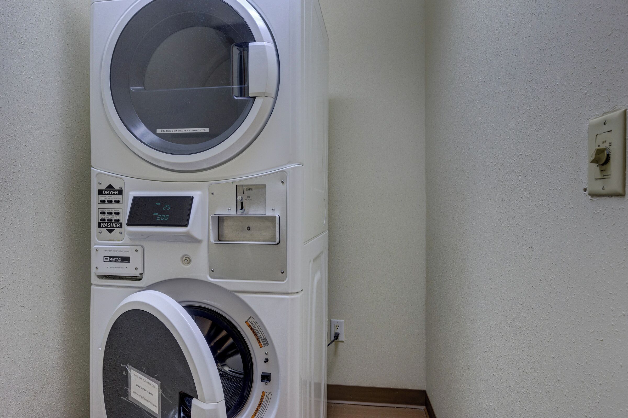 A stacked washer and dryer unit with a digital display, control panel, and a wall-mounted electrical outlet.