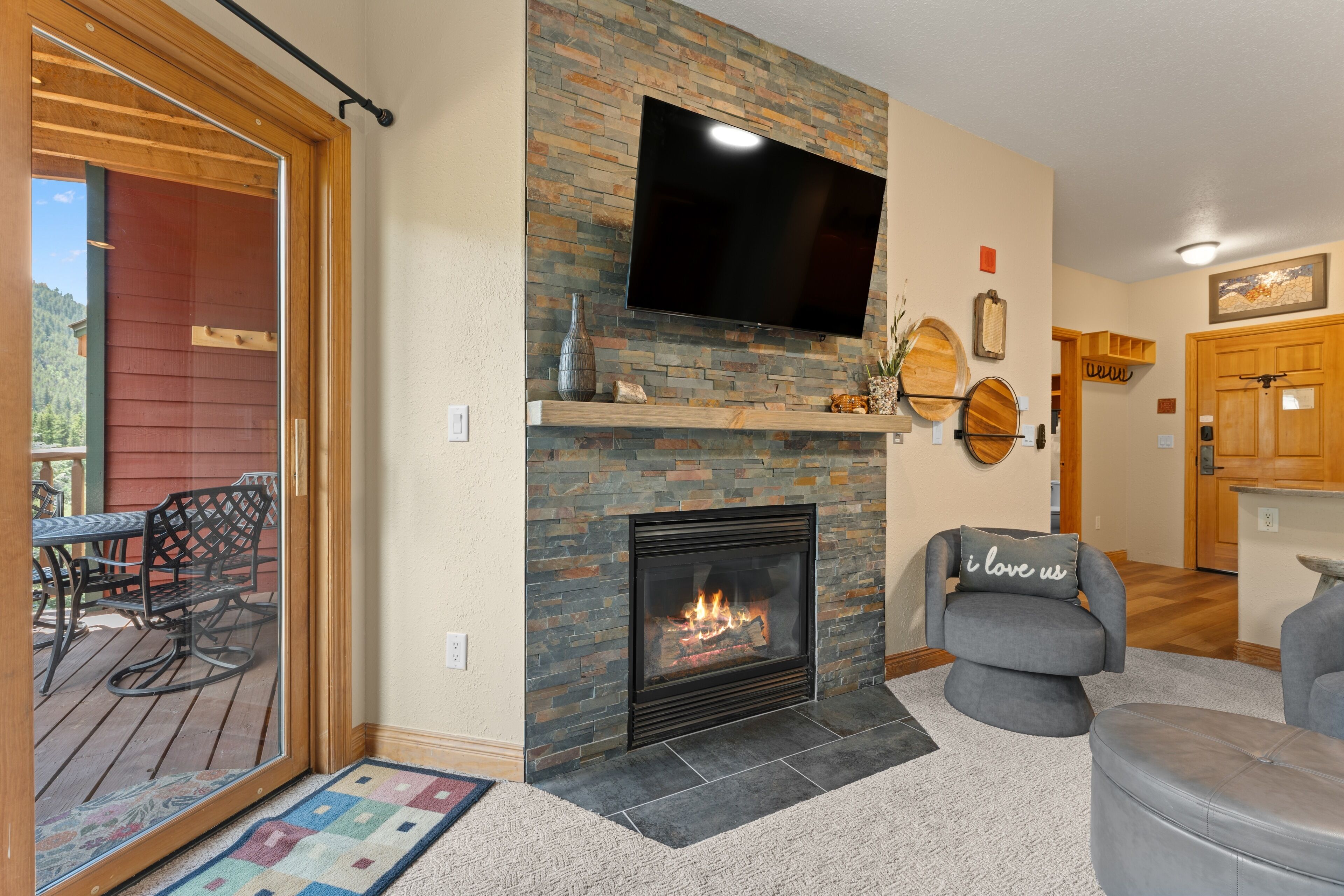 A living room with a stone fireplace, a flat-screen TV mounted above it, and a cozy seating area with a grey sofa and ottoman.