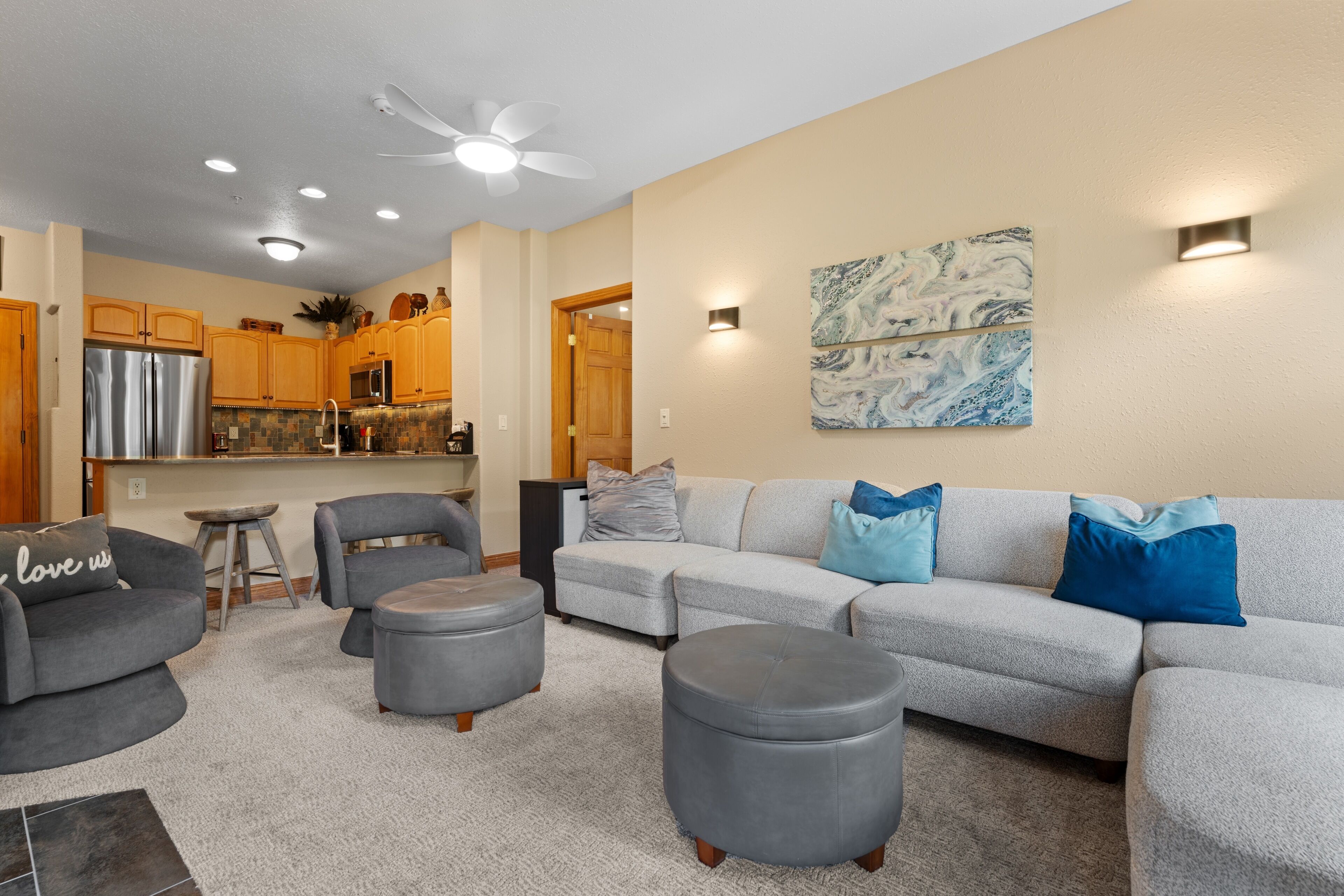 A modern living room with a grey sectional sofa, two ottomans, a grey armchair, and a coffee table. A ceiling fan and recessed lighting are visible.