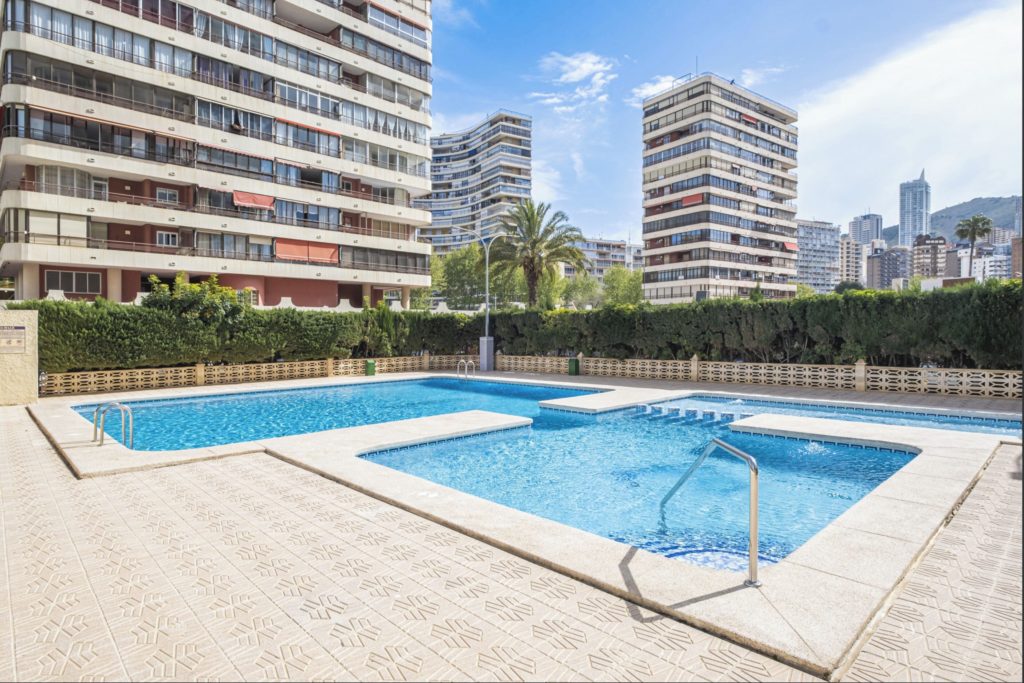 Zona de piscina con suelo de baldosas, rodeada por un seto y una valla de madera.