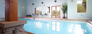 An indoor swimming pool with stone pillars, a seating area with tables and chairs, and large windows with a view of trees.