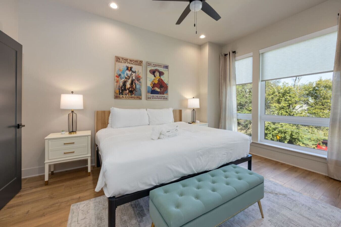 Quarto com cama, duas mesas de cabeceira, ventoinha no teto, janela com cortinas e cartazes nas paredes.