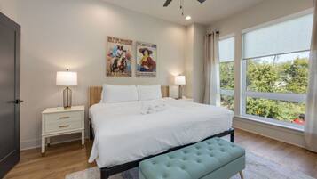 Quarto com cama, duas mesas de cabeceira, ventoinha no teto, janela com cortinas e cartazes nas paredes.