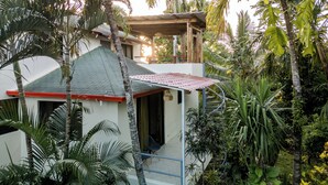A two-story house with a green roof, surrounded by lush tropical vegetation.