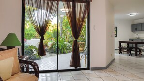A room with a sliding glass door leading to a garden, a dining table with chairs, and a sofa with cushions.