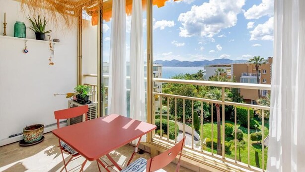 A balcony with a red table and chairs, a hanging plant, and a view of the sea and mountains. - Superb studio with sea and Esterel views, 2-minute walk to the beach (Cannes)