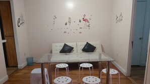 Une pièce avec une table en verre, des tabourets blancs, un canapé blanc avec des coussins noirs et des autocollants décoratifs sur les murs, représentant notamment un flamant rose et un chef cuisinier.