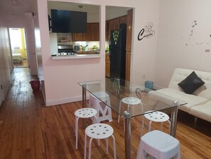 A modern kitchen with a TV, wooden cabinets, and a refrigerator. There is a glass dining table with stools.