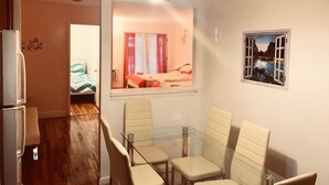 Une pièce comprenant une table à manger en verre, des chaises, un réfrigérateur, un lit avec des rideaux rouges et un tableau représentant un paysage.