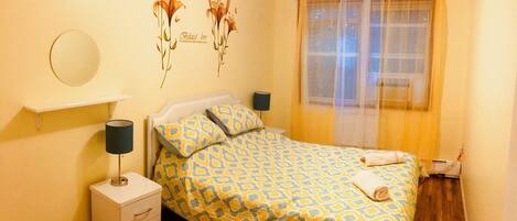 A bedroom with a bed, a bedside table, a lamp, a wall decoration, a window with curtains, and a small trash bin.