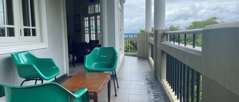 A balcony with a wooden table and two green chairs, a white railing, and a view of the sea.