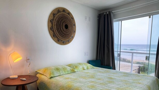 Una camera da letto con un letto, una lampada da comodino, un addobbo a parete, una finestra con tende e vista sull'oceano.