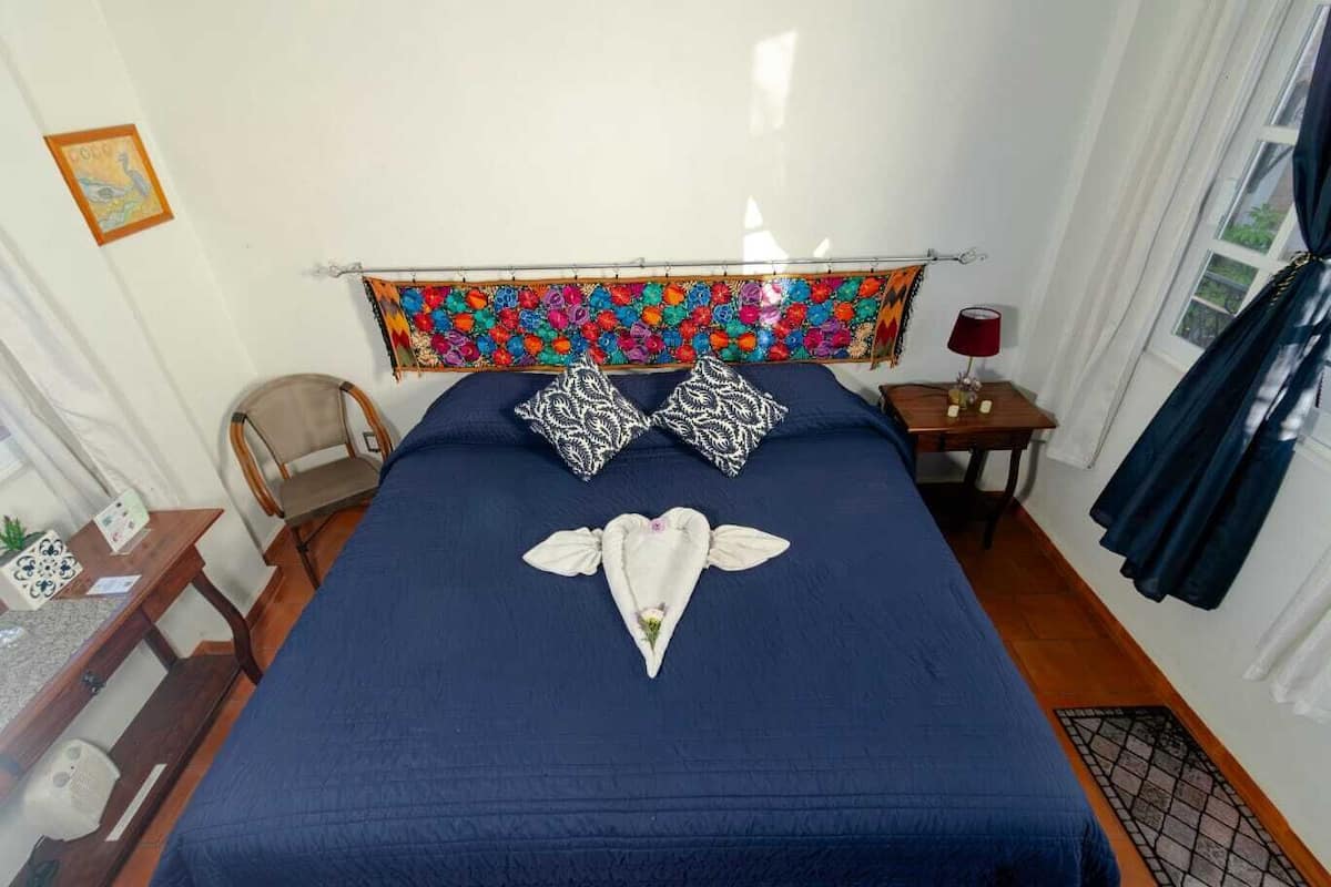 A neatly made bed with a blue bedspread and two patterned pillows, a small bedside table with a lamp, and a chair in a room with white walls and a colorful tapestry above the bed.