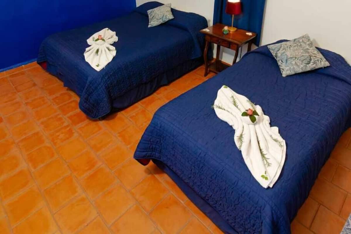 A hotel room with two beds, a blue wall, a red curtain, and a small table with a lamp.