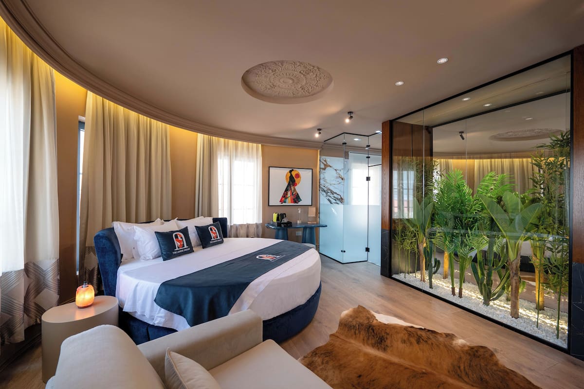 A modern hotel room with a large bed, a glass partition, and a view of indoor plants.