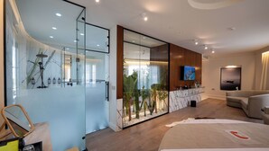 A modern hotel room with a glass shower enclosure, a large mirror, a wooden vanity, a flat-screen TV, a sofa, and a bed with a red telephone symbol. - The Collector hotel boutique (Sarandë)