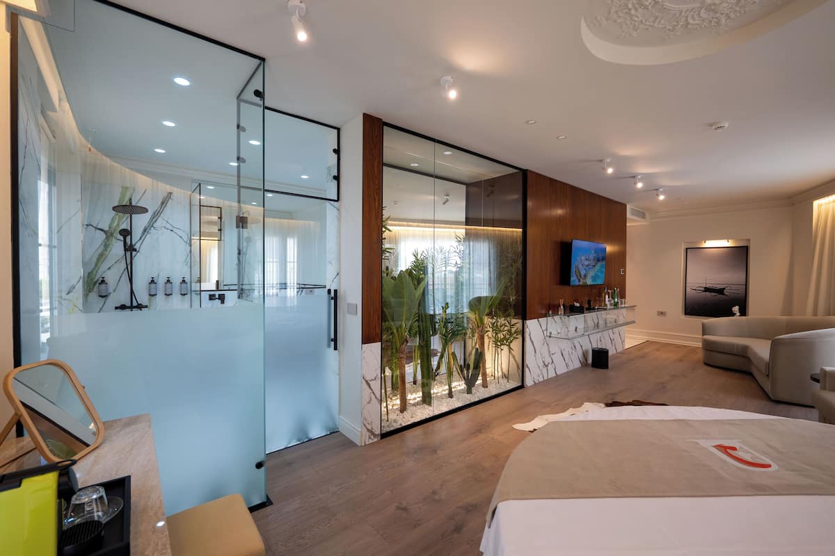 A modern hotel room with a glass shower enclosure, a large mirror, a wooden vanity, a flat-screen TV, a sofa, and a bed with a red telephone symbol.