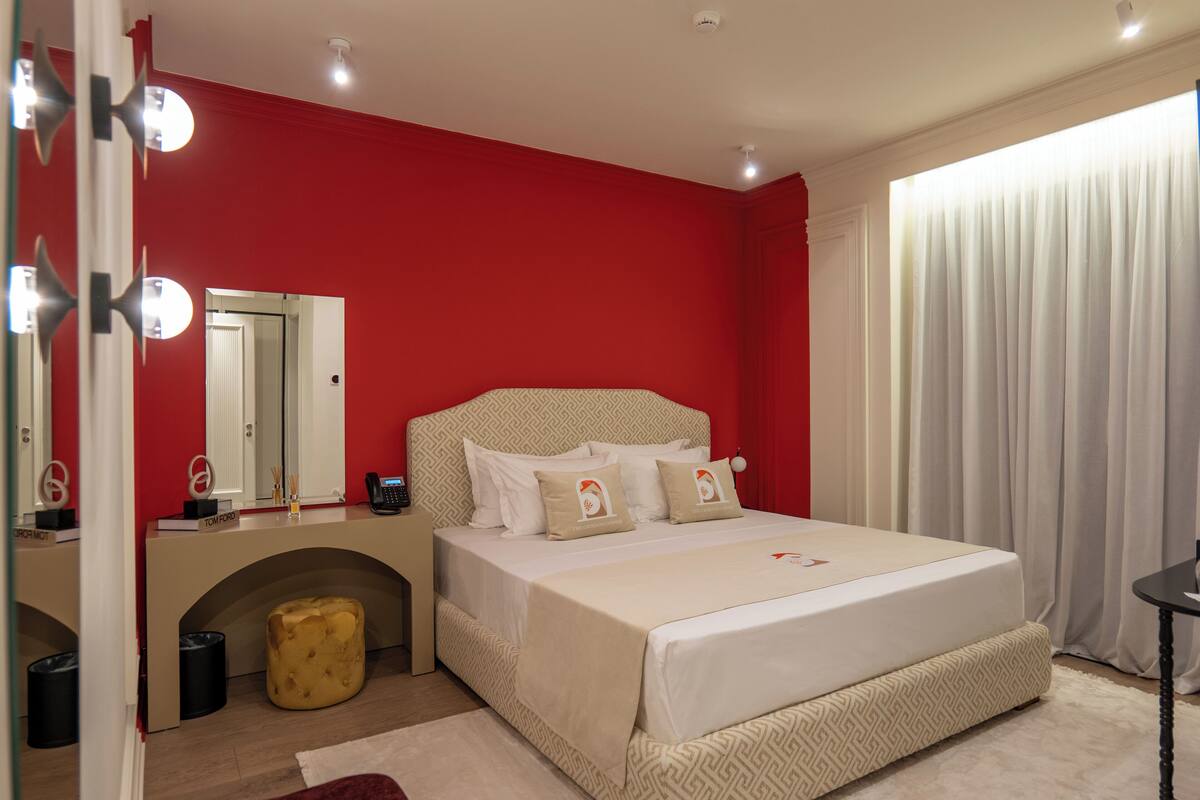 A bedroom with a red wall, a bed with white bedding, a vanity with a mirror, and a window with curtains.