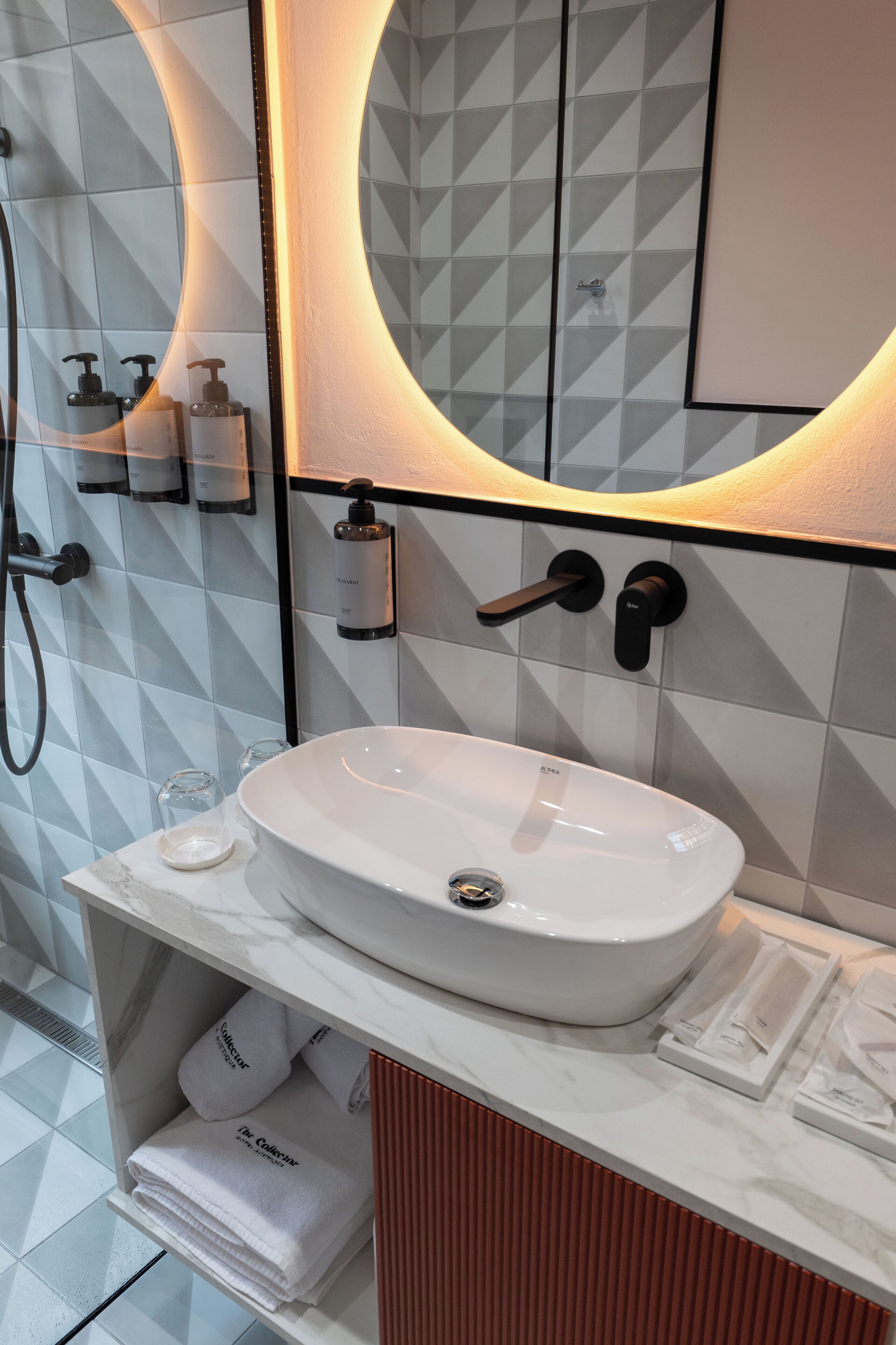 A modern bathroom with a white sink, a mirror with a circular light, and a shower area.