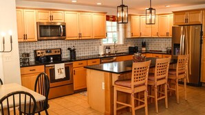 A kitchen with wooden cabinets, a stainless steel refrigerator, a microwave, and a coffee maker. There are three wooden chairs around a central island. - Large Cozy Home •Songo River•Brandy Pond•Long Lake•Sebago (Naples)