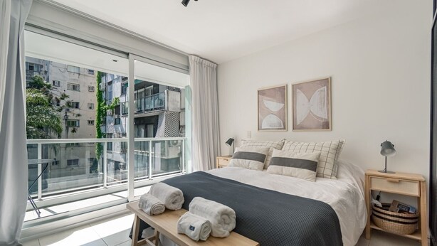 A modern bedroom with a large bed, two bedside tables, a bench with towels, and a view of the city through a large window. - Comfortable stylish studio in Recoleta (Ciudad Autonoma de Buenos Aires)