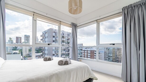 A modern hotel room with a large bed, two windows, and a city view.