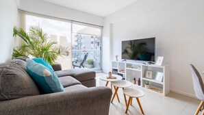 A modern living room with a grey sofa, teal pillow, two small white tables, a TV on a white stand, and a balcony with a view of buildings.