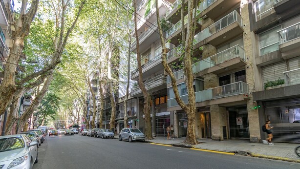 A multi-story residential building with balconies, parked cars, and trees lining the street.