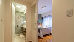 Ein Flur, der zu einem Badezimmer mit Dusche und Toilette sowie zu einem Schlafzimmer mit Bett und Vorhängen führt, mit Holzboden.