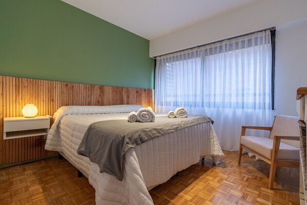 A hotel room with a wooden headboard, a bed with white bedding, a chair, and a window with sheer curtains. - Spacious apartment in Palermo (caba)