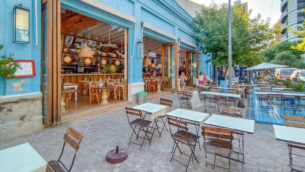 Ein Straßencafé mit Tischen und Stühlen, ein blaues Gebäude mit offener Eingangstür und eine Straße mit Autos und Bäumen.