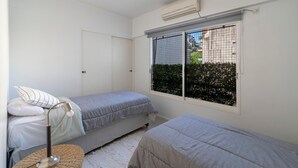 Ein kleines, sauberes Zimmer mit zwei Einzelbetten, einer Nachttischlampe, einem Fenster mit Blick auf Grünflächen und einer Klimaanlage.