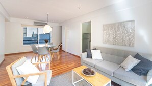 Ein modernes Wohnzimmer mit grauem Sofa, einem Esstisch mit Stühlen, einem Holztisch und einem großen Fenster mit Blick auf die Stadt.