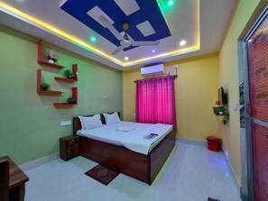 A bedroom with a bed, nightstands, a ceiling fan, a red trash bin, and a window with pink curtains. - SAMRIDDHA BEACH RESORT (Kanthi)