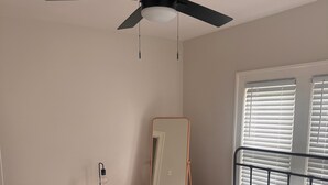 A bedroom with a bed, a ceiling fan, a bedside table with a lamp, a mirror, and a window with blinds.