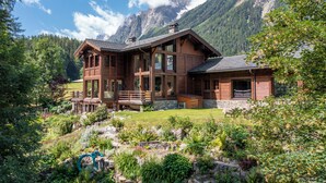A large wooden chalet with multiple levels, a stone foundation, and a balcony overlooking a lush garden.